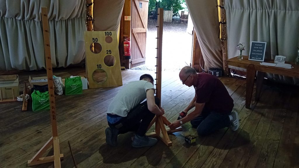 A black and white image showing two men crouched on a wooden floor inside a stretch tent, as they build a wooden Limbo stand. In the background the wooden garden games stand ready near the exit.