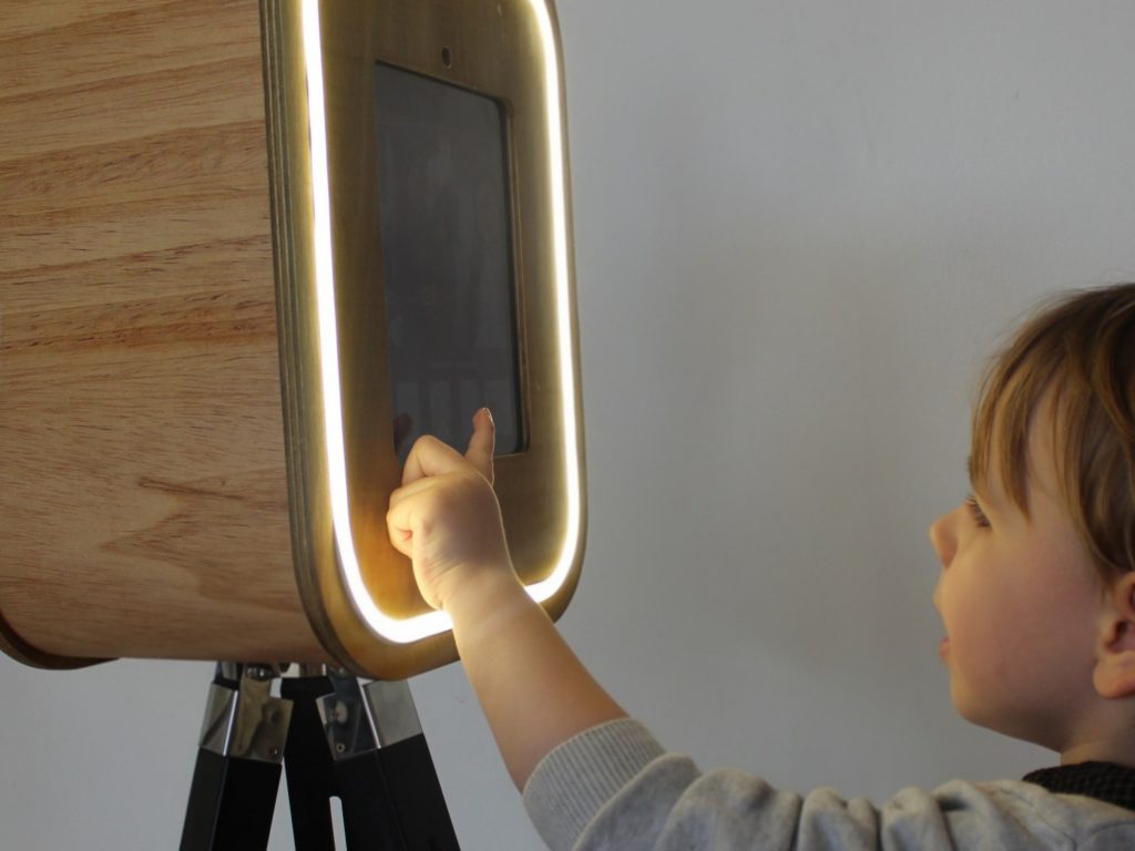 Close-up of a wooden photobooth in use, showing a child's hand interacting with the screen, with a cropped side profile visible on the right.